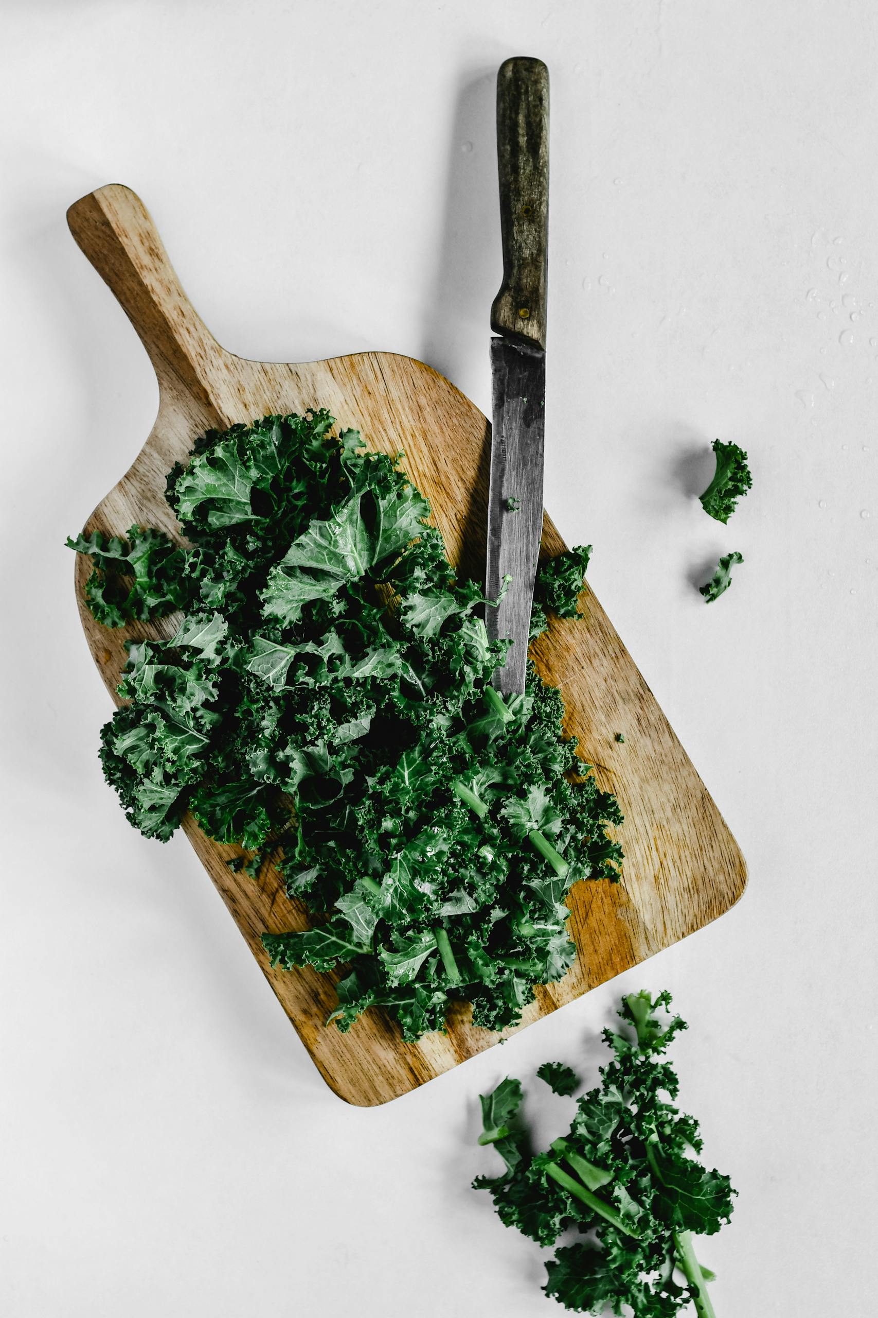best foods for hormone balance: Overhead view of fresh kale on a wooden board with a knife on a white surface.