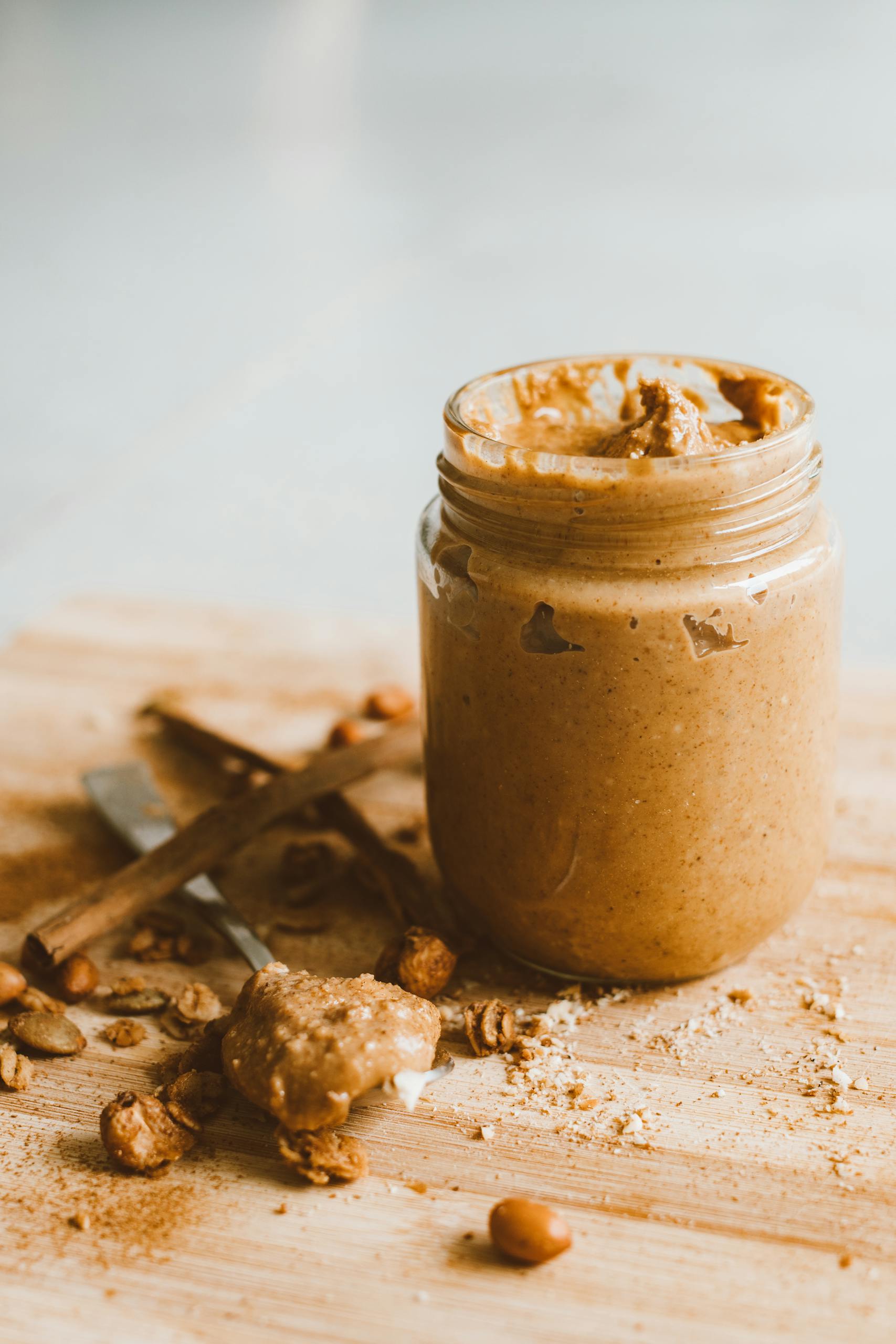 high-protein vegan snacks: A jar of creamy peanut butter with scattered peanuts and spoon on a wooden surface.