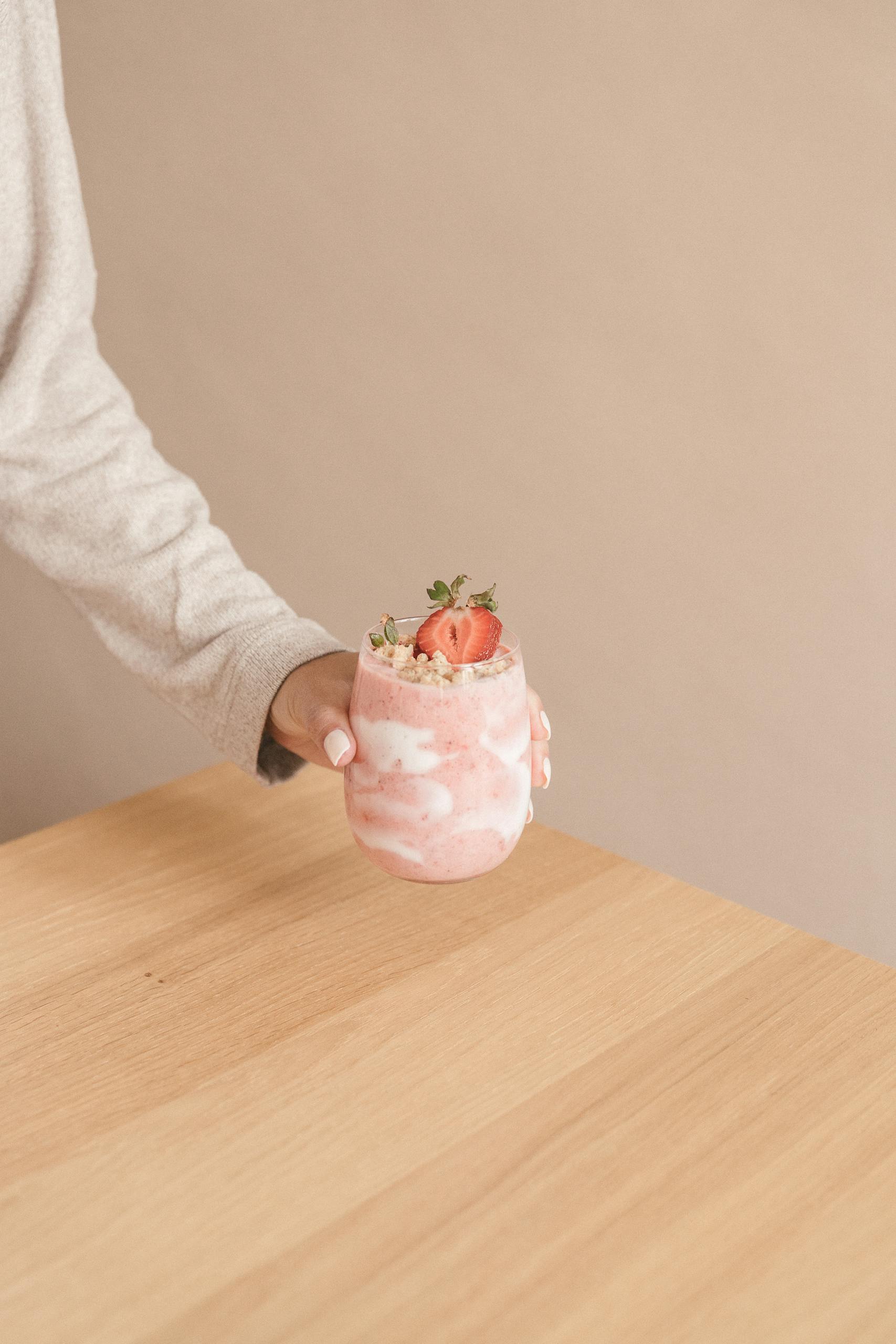 kefir benefits: A hand holding a pink strawberry smoothie in a glass on a wooden table, against a soft background.