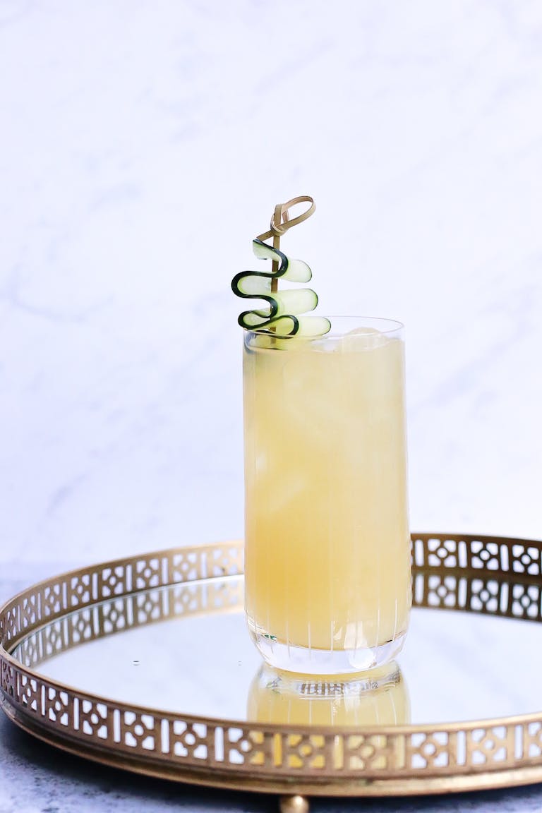 clear protein drinks: A tall glass of refreshing citrus beverage garnished with a cucumber spiral on a decorative tray.