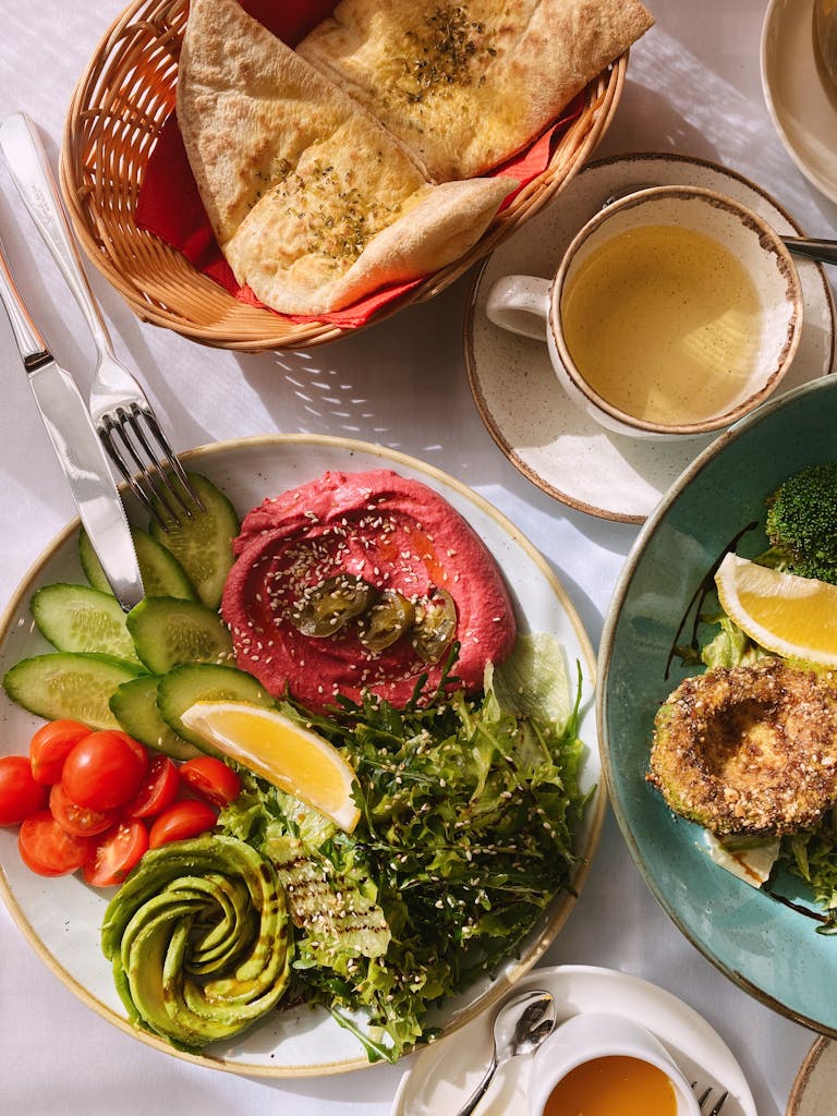 lipedema diet: A vibrant vegan brunch featuring hummus, fresh veggies, tea, and pita bread on a sunlit table.