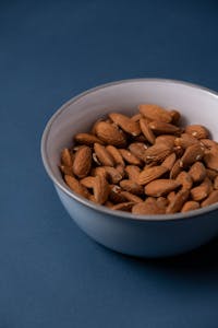 simple ways to increase calcium intake: Close-up of raw almonds in a ceramic bowl against a blue backdrop, showcasing healthy nutrition.
