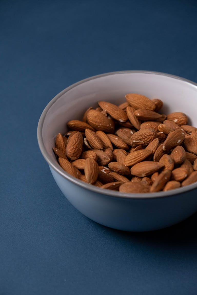 simple ways to increase calcium intake: Close-up of raw almonds in a ceramic bowl against a blue backdrop, showcasing healthy nutrition.