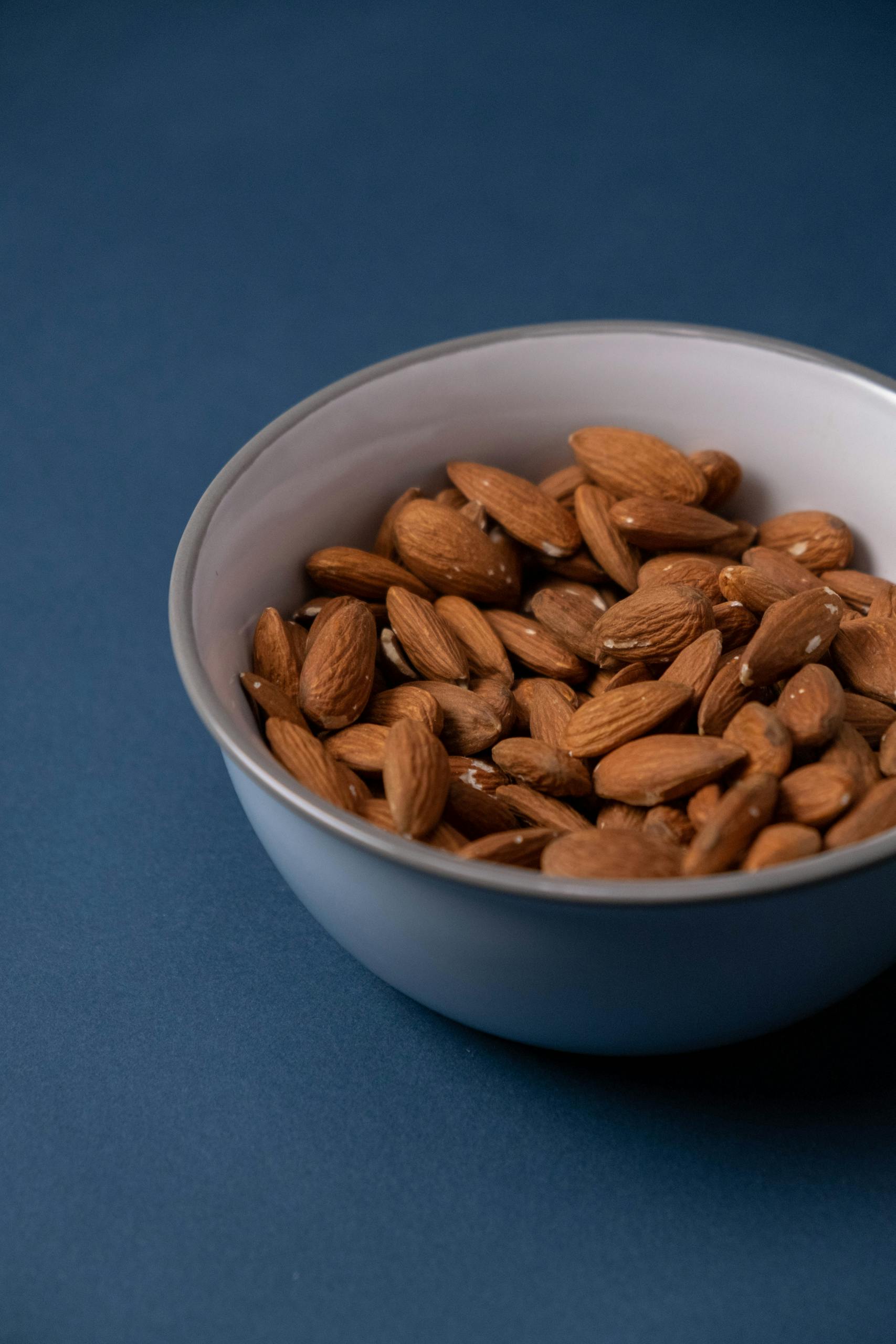 simple ways to increase calcium intake: Close-up of raw almonds in a ceramic bowl against a blue backdrop, showcasing healthy nutrition.