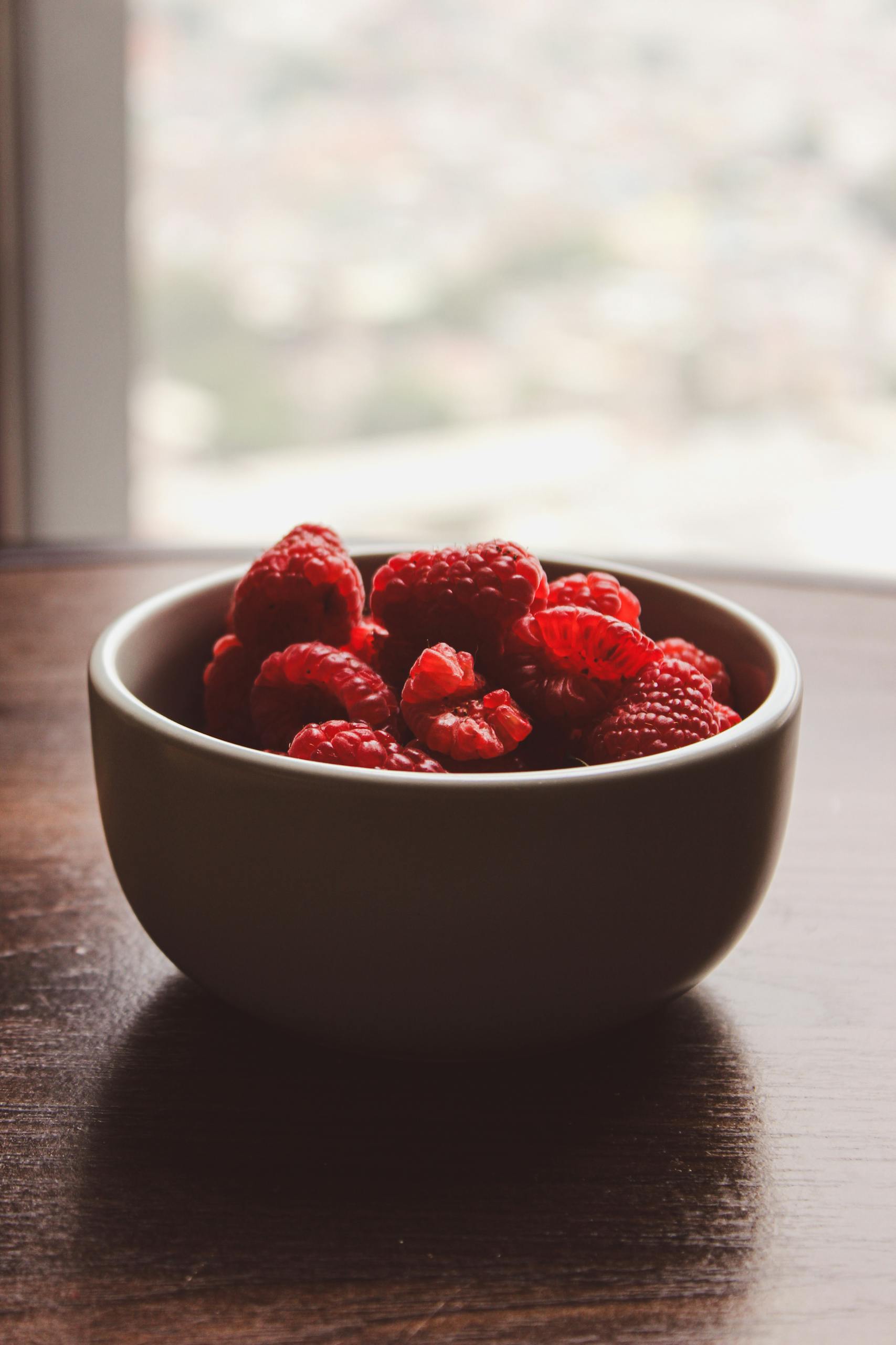 GLP-1 diet: Close-up of vibrant red raspberries in a bowl on a wooden table by a window.