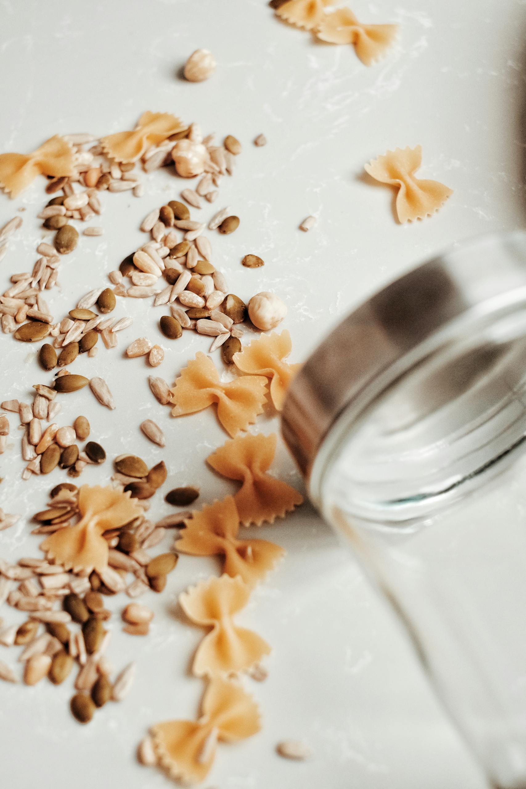 fibermaxxing: Delicate bow-tie pasta and assorted seeds artistically scattered near a glass jar on a table.