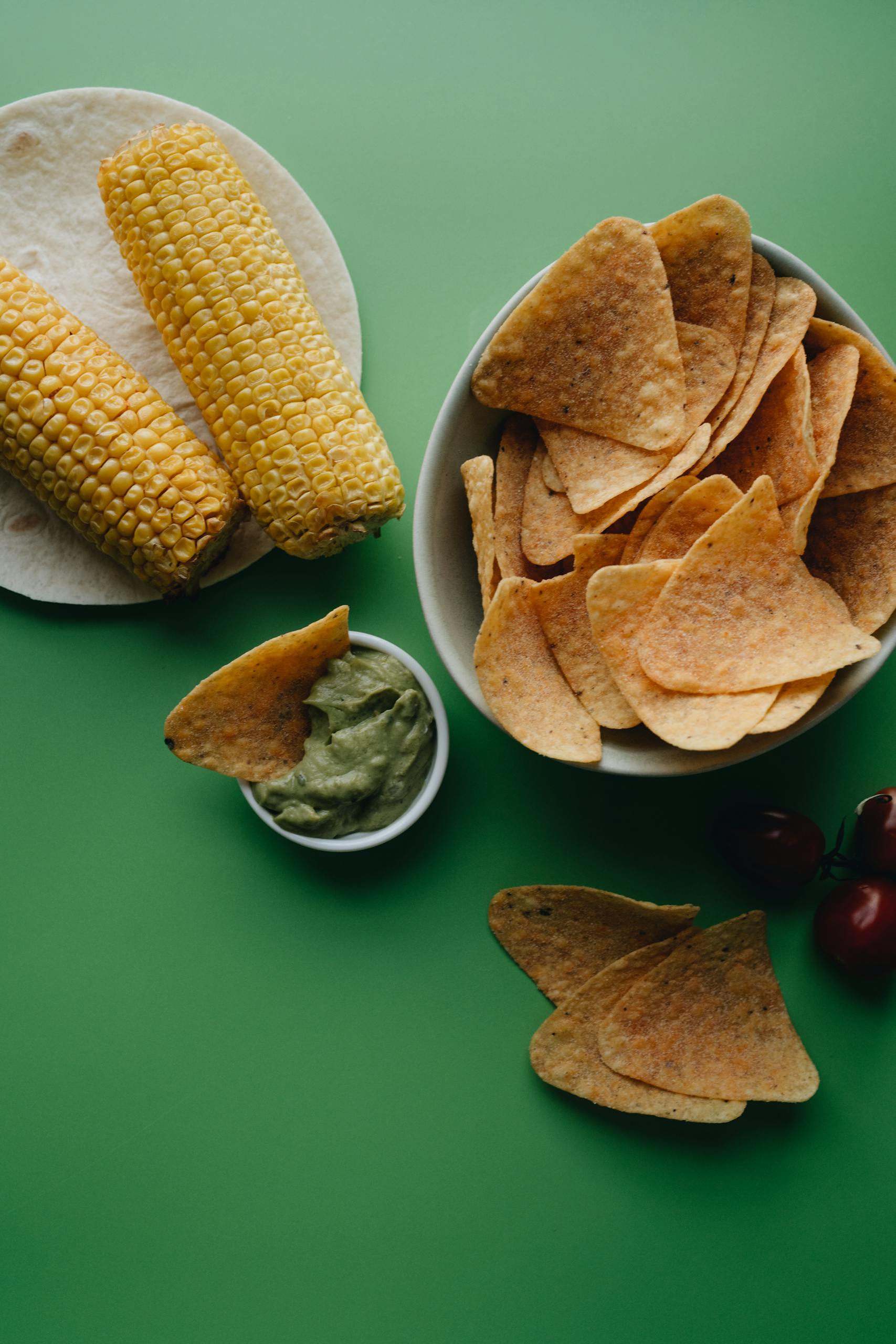 Corn Tortillas vs. Flour Tortillas: Flat lay of Mexican snack foods including tortilla chips, guacamole, and corn on a green background.