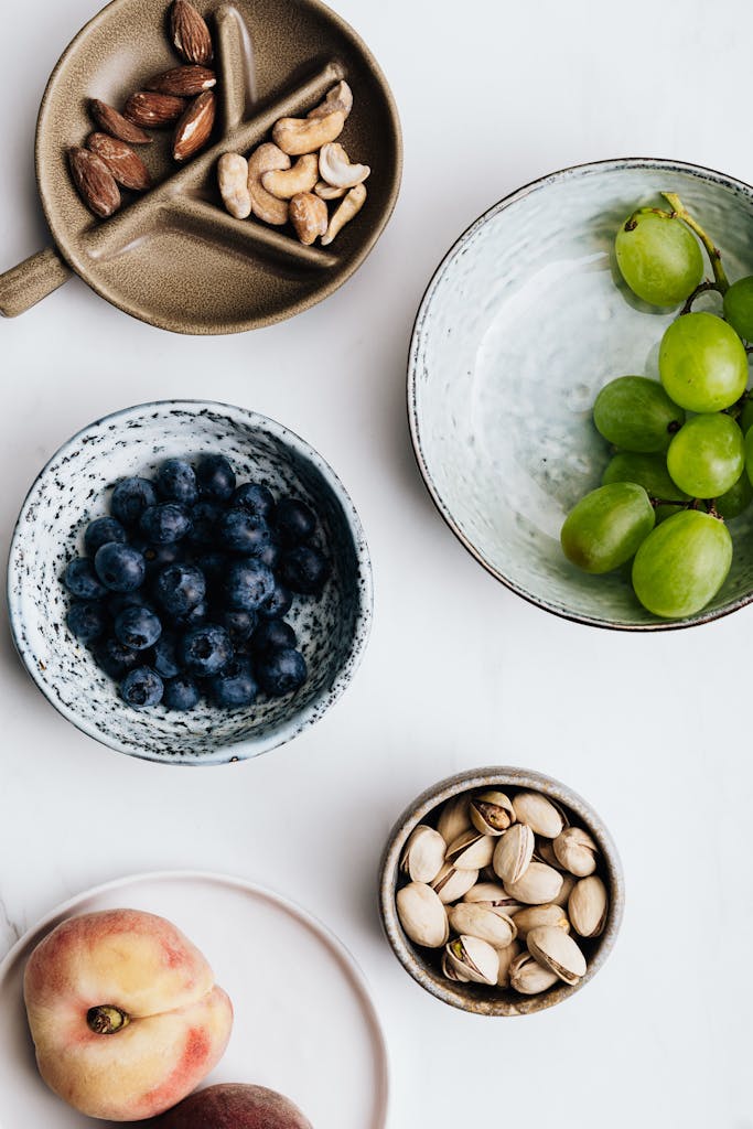 best healthy snacks at whole foods market: Flatlay of nuts, fruits, and berries in bowls on a white background offers a healthy snack choice.