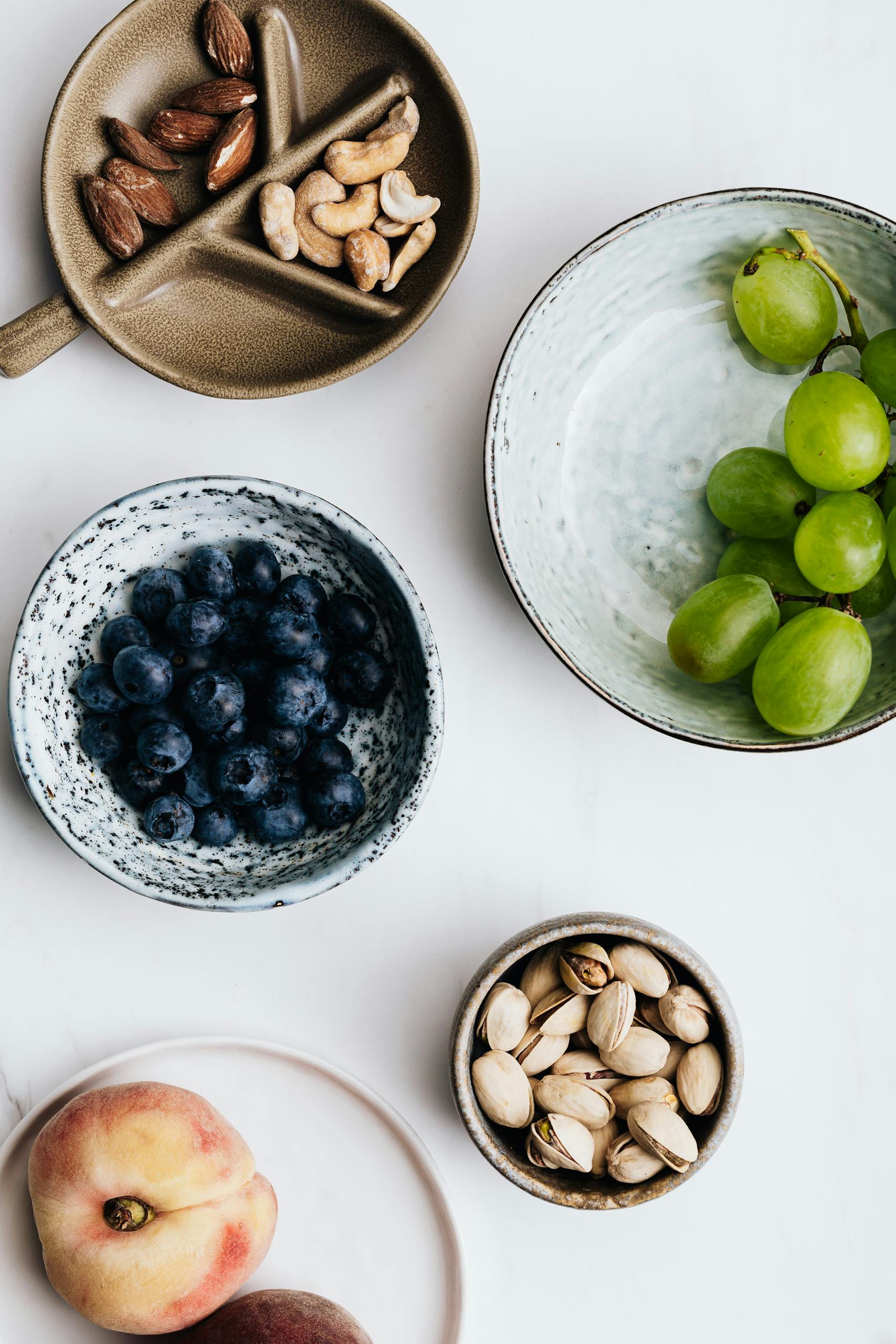 best healthy snacks at whole foods market: Flatlay of nuts, fruits, and berries in bowls on a white background offers a healthy snack choice.