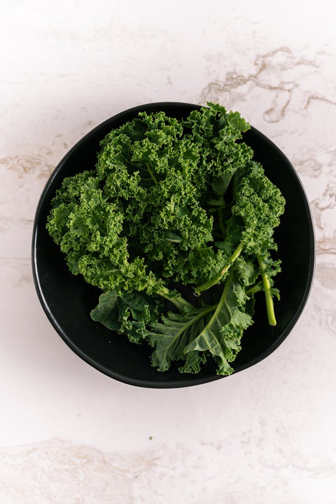 spinach vs kale nutrition: Top view of fresh kale leaves in a black bowl against a marble background. Perfect for healthy eating visuals.