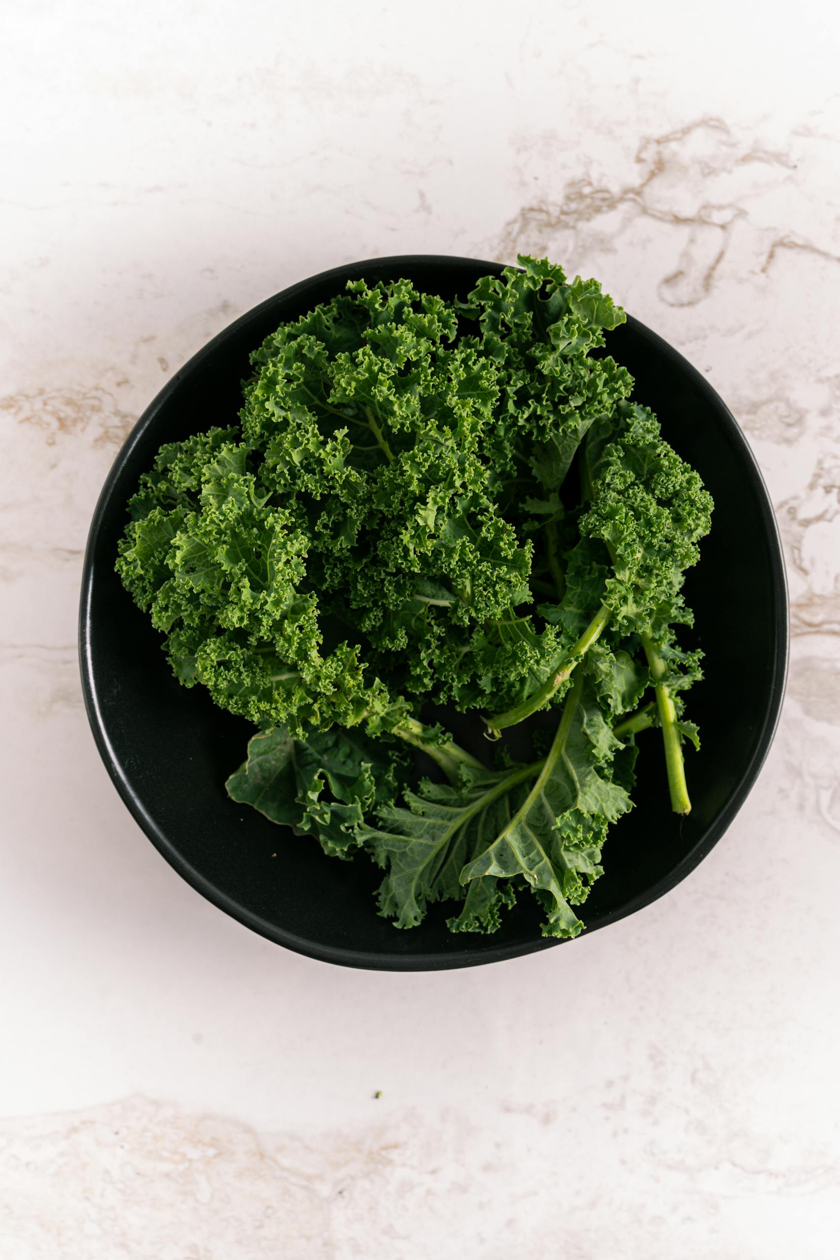 spinach vs kale nutrition: Top view of fresh kale leaves in a black bowl against a marble background. Perfect for healthy eating visuals.