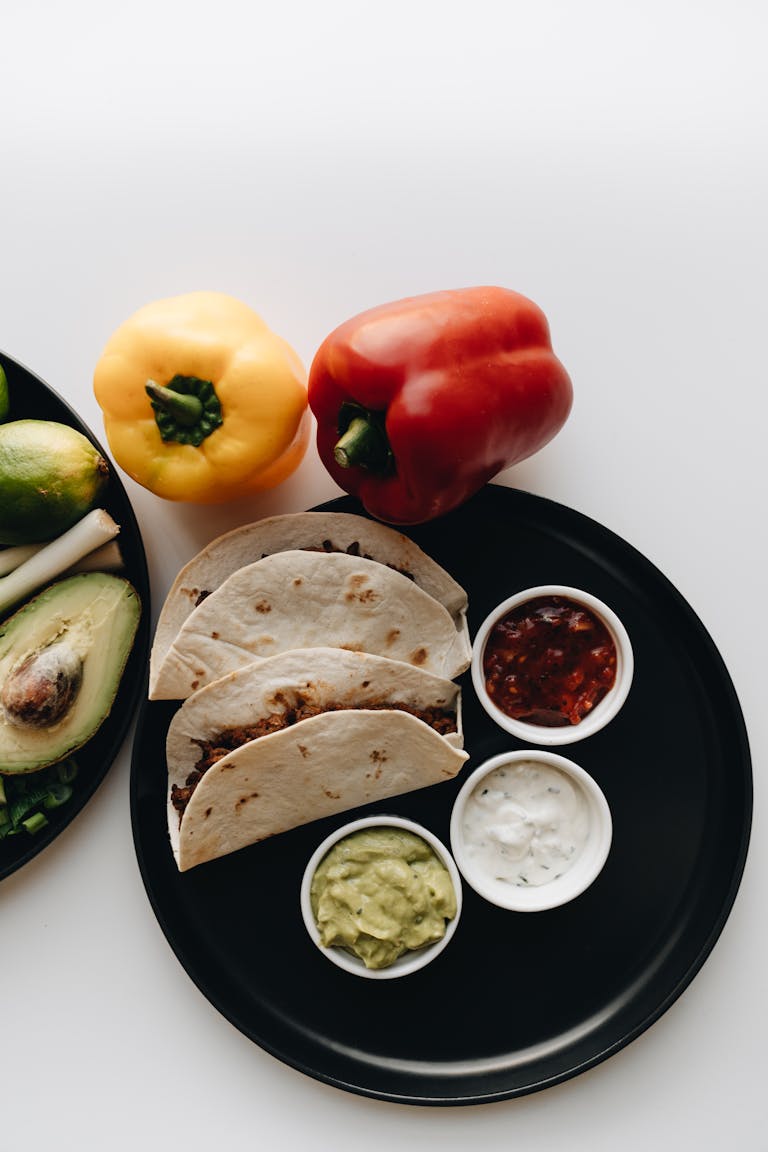 Corn Tortillas vs. Flour Tortillas: Top view of tacos with salsa, bell peppers, and avocado on a black plate on a white surface.