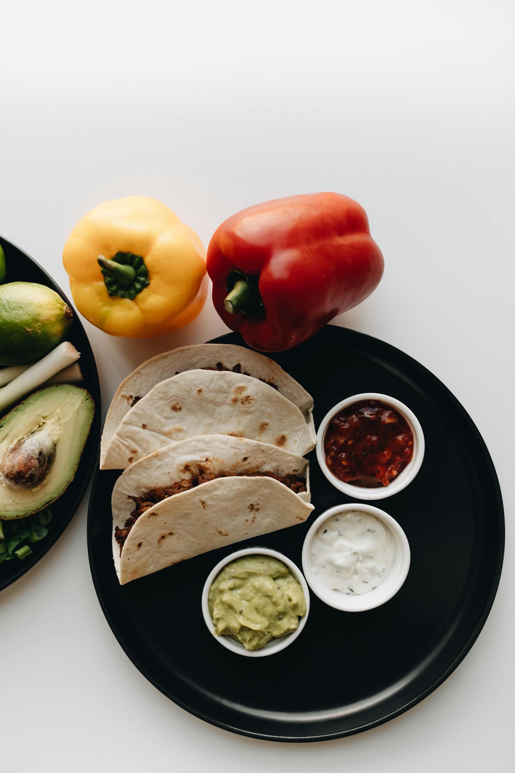 Corn Tortillas vs. Flour Tortillas: Top view of tacos with salsa, bell peppers, and avocado on a black plate on a white surface.