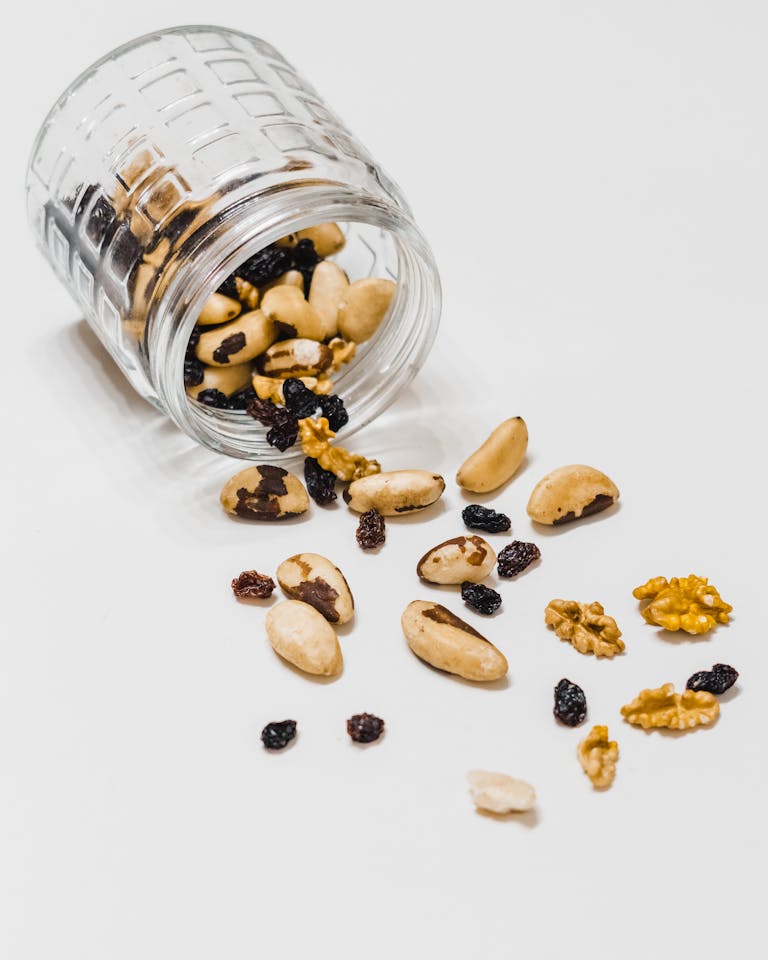is snacking bad for metabolism: A glass jar spills assorted nuts and raisins on a white surface, perfect for snack themes.