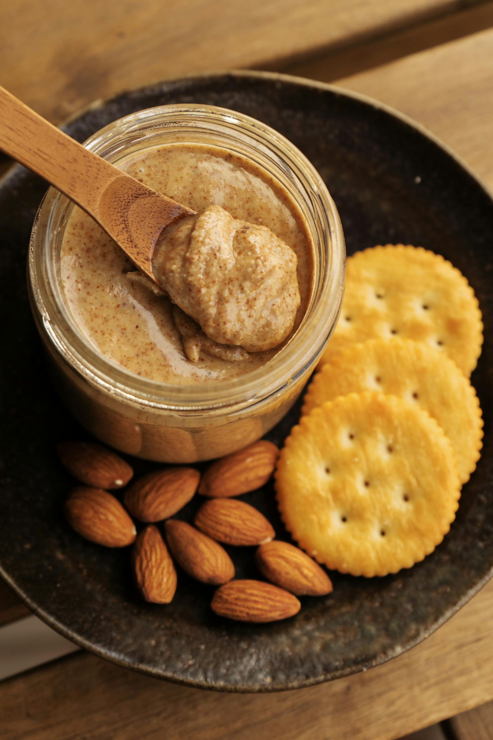 Healthy Fats for Breakfast: A jar of creamy almond butter with a wooden spoon, surrounded by almonds and crackers, offering a healthy snack option.
