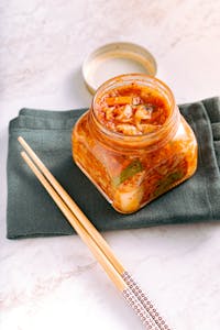 probiotic foods vs. supplements: A jar of traditional homemade kimchi on a marble surface with wooden chopsticks beside it.