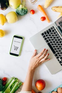 How to Shop Healthy on a Budget: A top view of online grocery shopping using a laptop and smartphone surrounded by fresh vegetables and fruits.