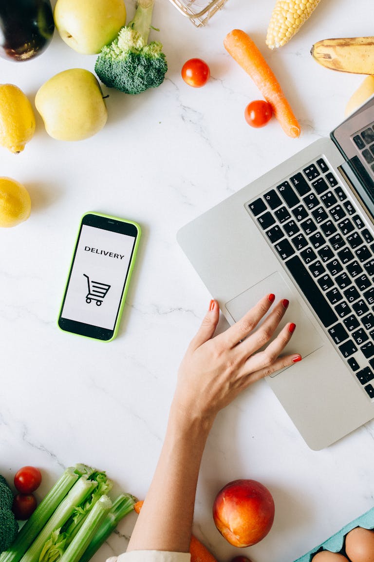 How to Shop Healthy on a Budget: A top view of online grocery shopping using a laptop and smartphone surrounded by fresh vegetables and fruits.
