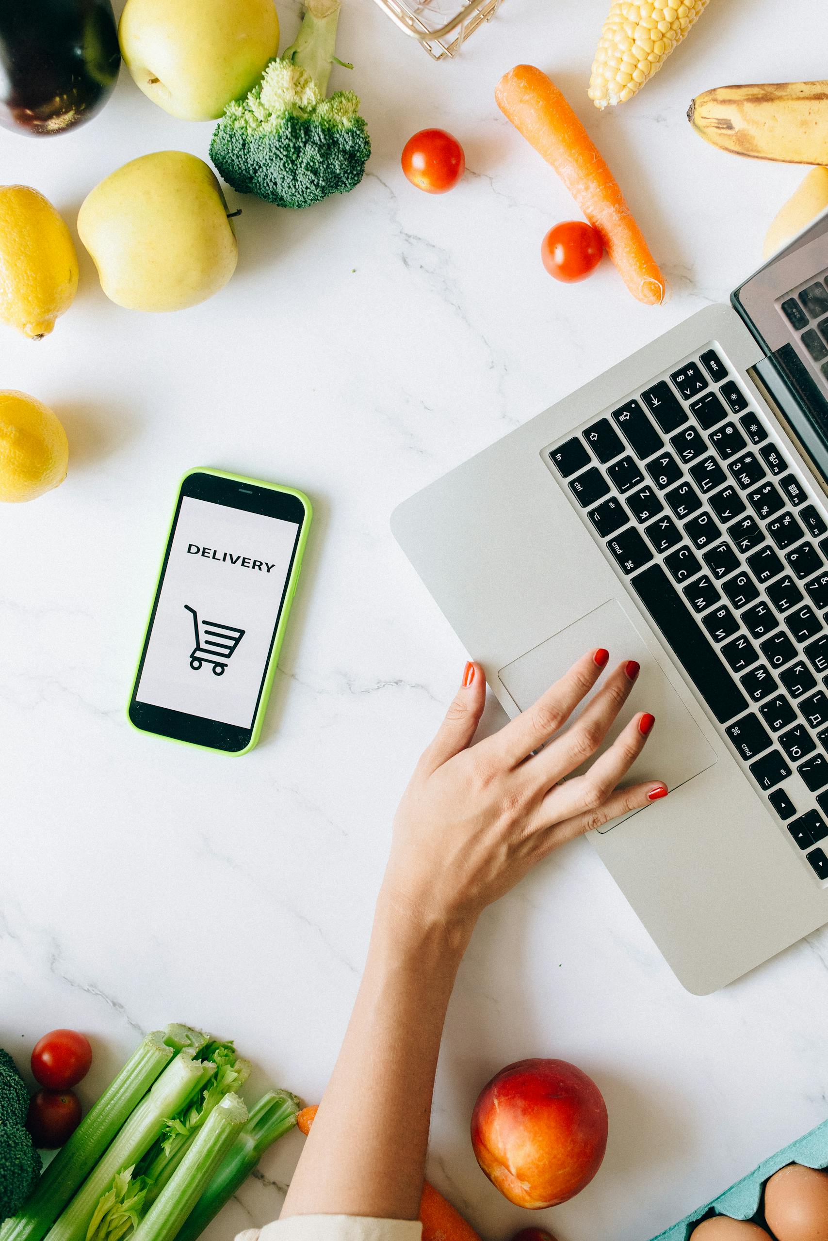 How to Shop Healthy on a Budget: A top view of online grocery shopping using a laptop and smartphone surrounded by fresh vegetables and fruits.