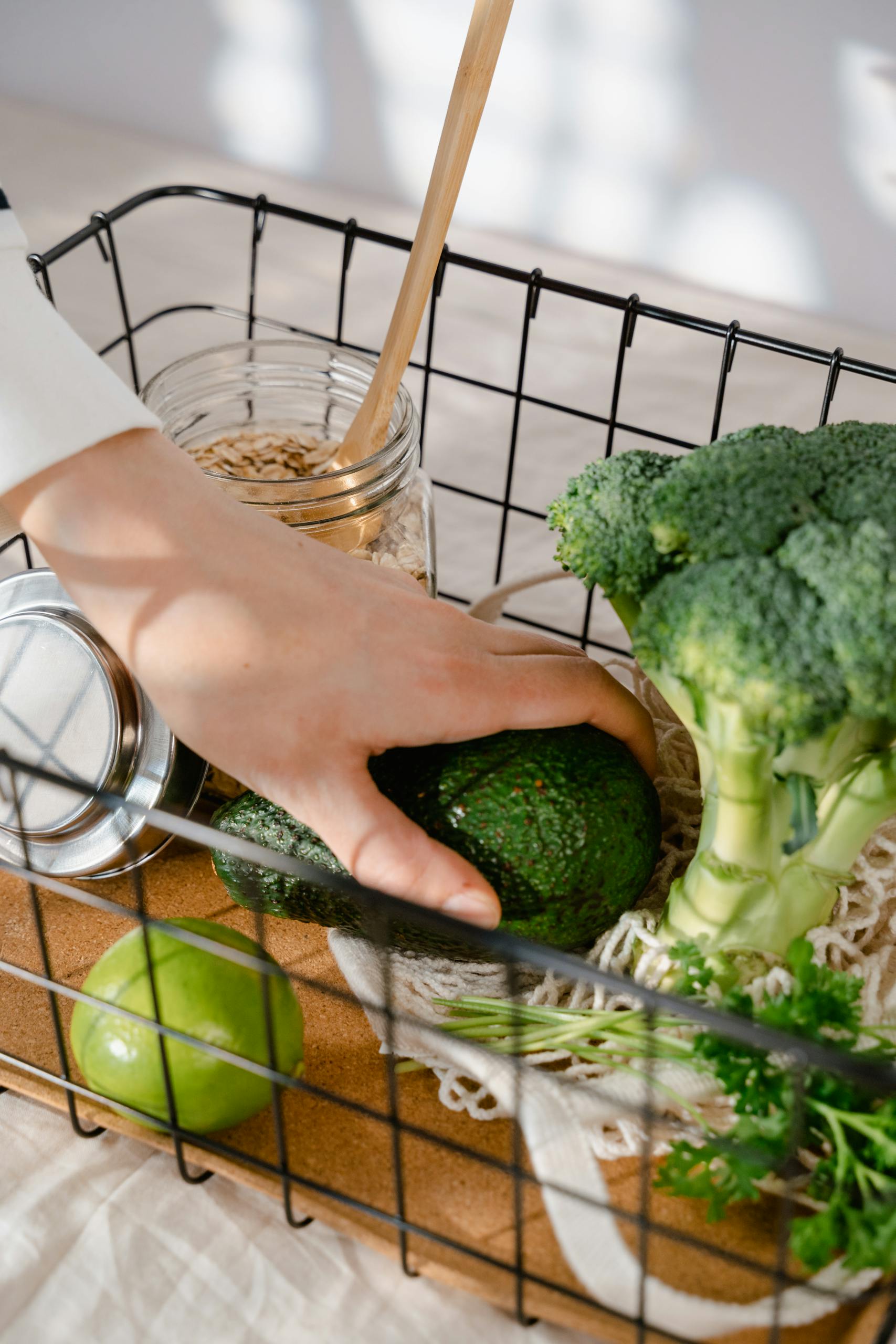 How to Shop Healthy on a Budget: Hand reaches for avocado amidst broccoli and grains in wire basket for healthy cooking.