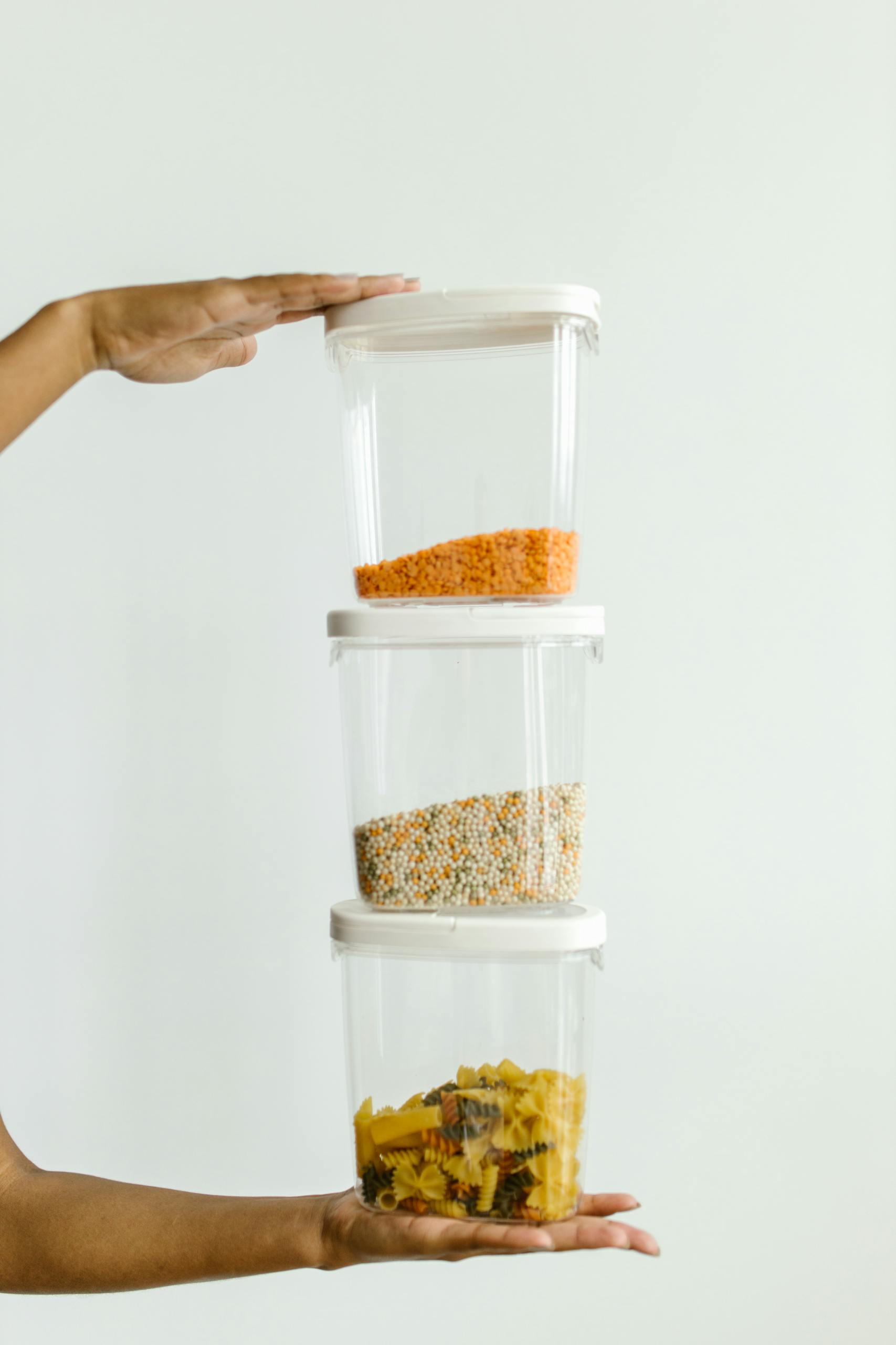 How to Reduce Food Waste at Home: Hands holding stacked plastic containers with various dry foods against a neutral background.