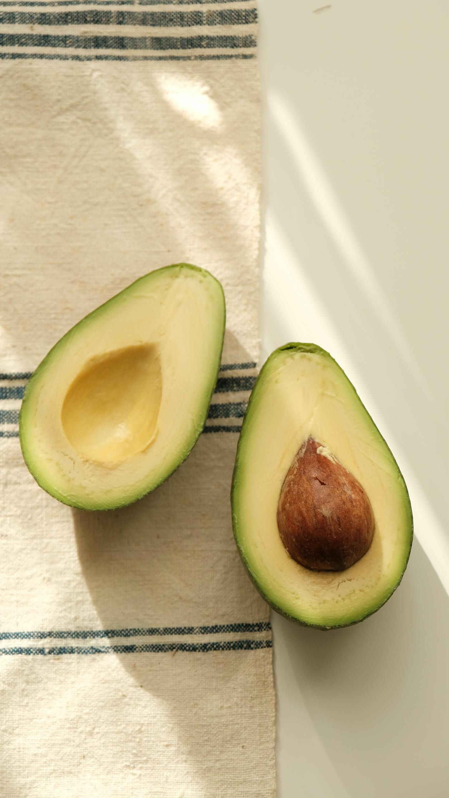 olive oil vs. avocado oil: Sliced ripe avocado halves with pit on a rustic striped cloth, sunlight casts gentle shadows.