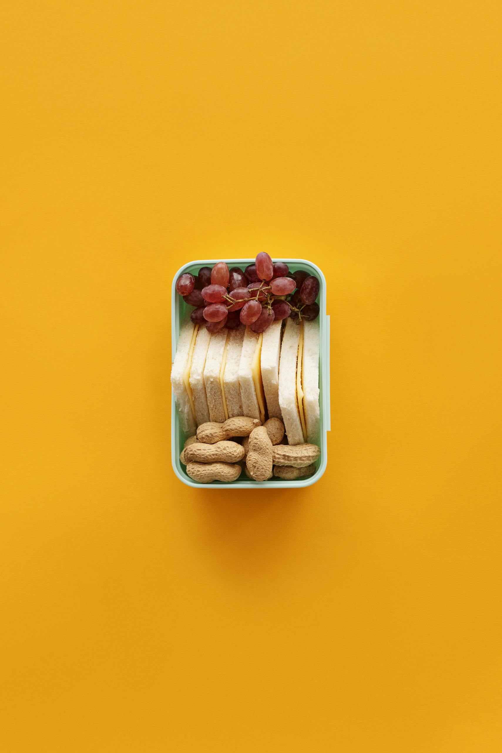 Best Post-Workout Meals for Muscle Recovery: Top view of a lunch box with grapes, sandwich, and peanuts on yellow background.