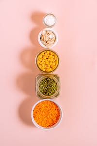high-protein, high-fiber foods: Vertical arrangement of various seeds and grains in bowls on a pink background.