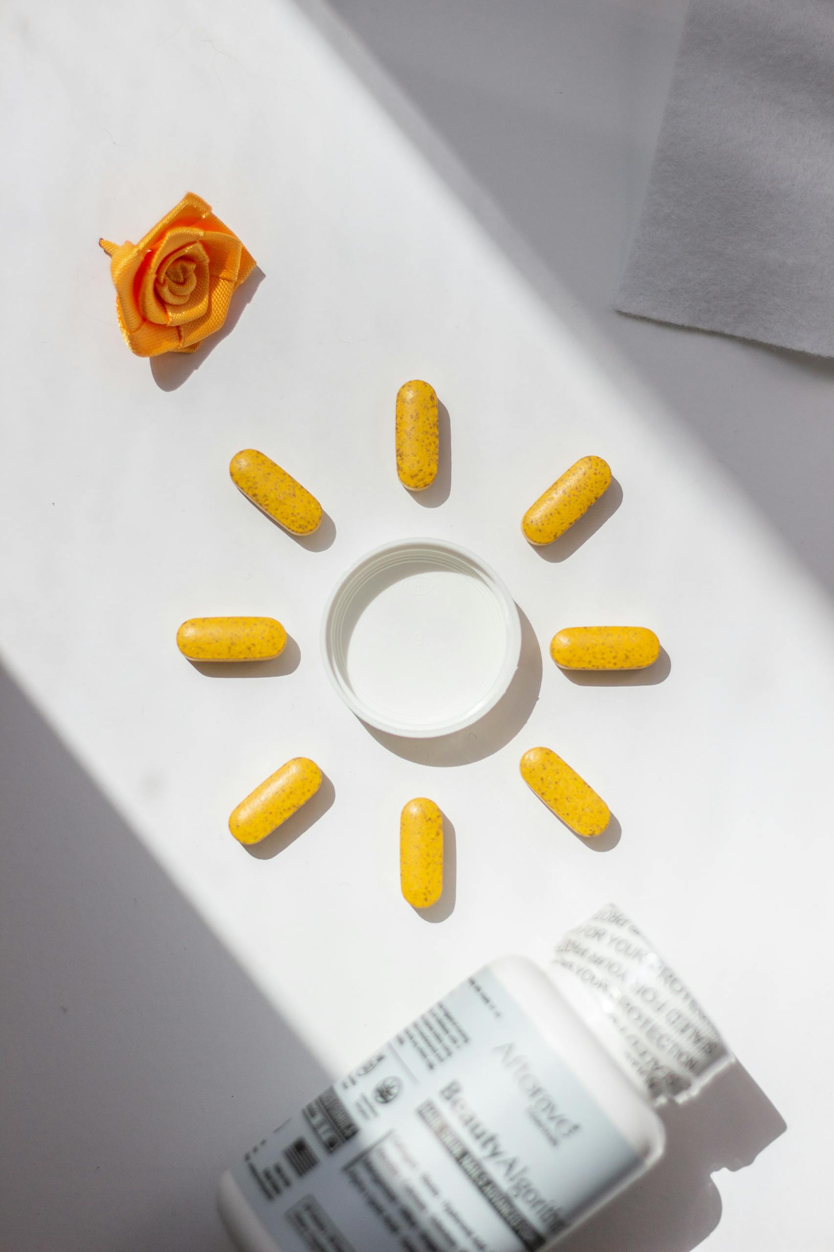 do you need vitamin D supplements: Bright yellow vitamin pills arranged in a sun shape with bottle and rose accent on white background.