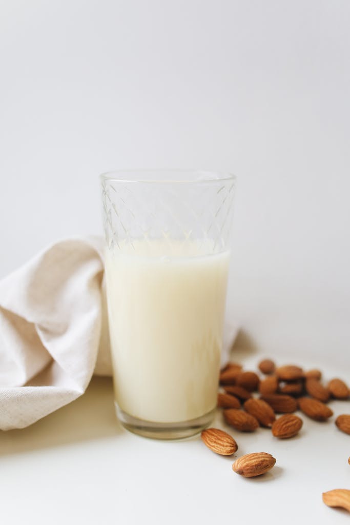 oat milk vs. almond milk vs. soy milk: Close-up of a glass of almond milk surrounded by almonds on a white surface, emphasizing vegan and plant-based lifestyle.