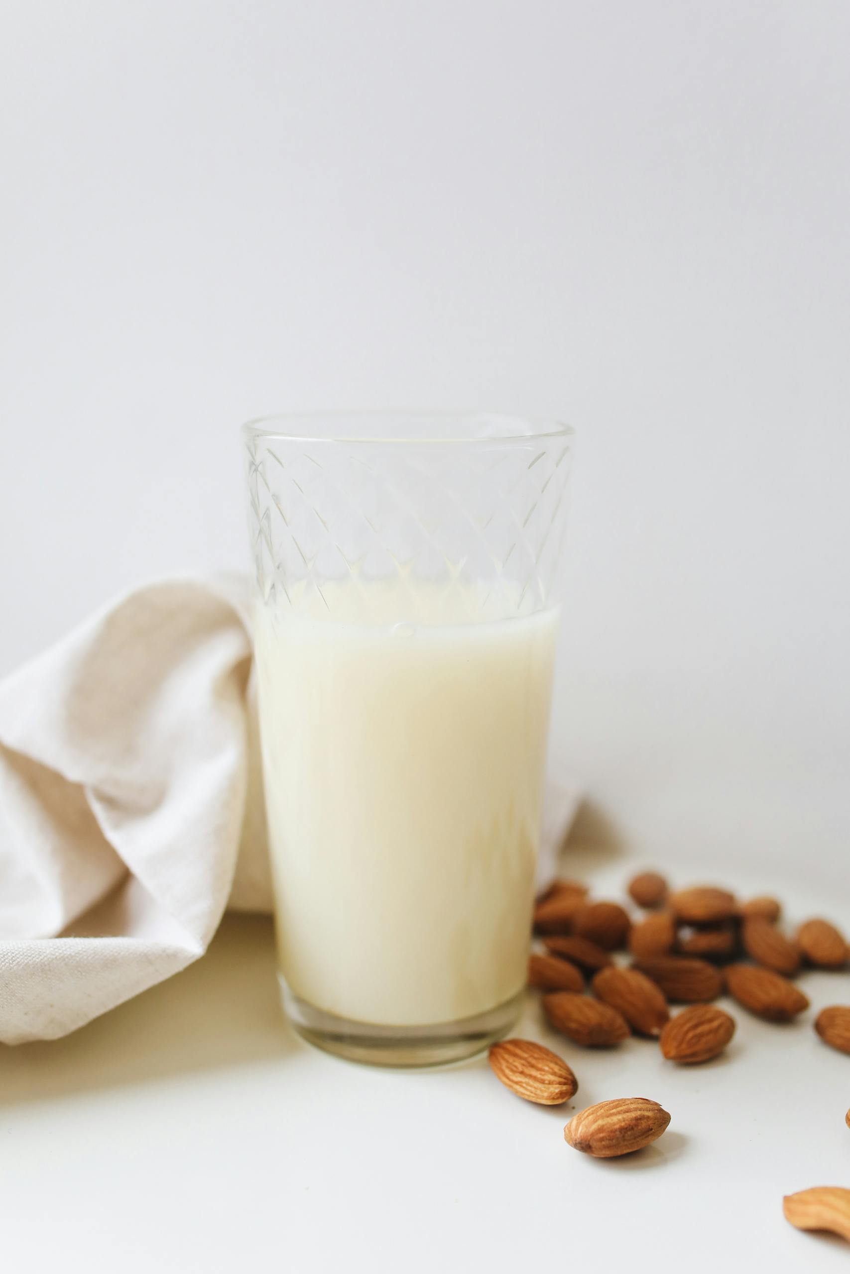 oat milk vs. almond milk vs. soy milk: Close-up of a glass of almond milk surrounded by almonds on a white surface, emphasizing vegan and plant-based lifestyle.