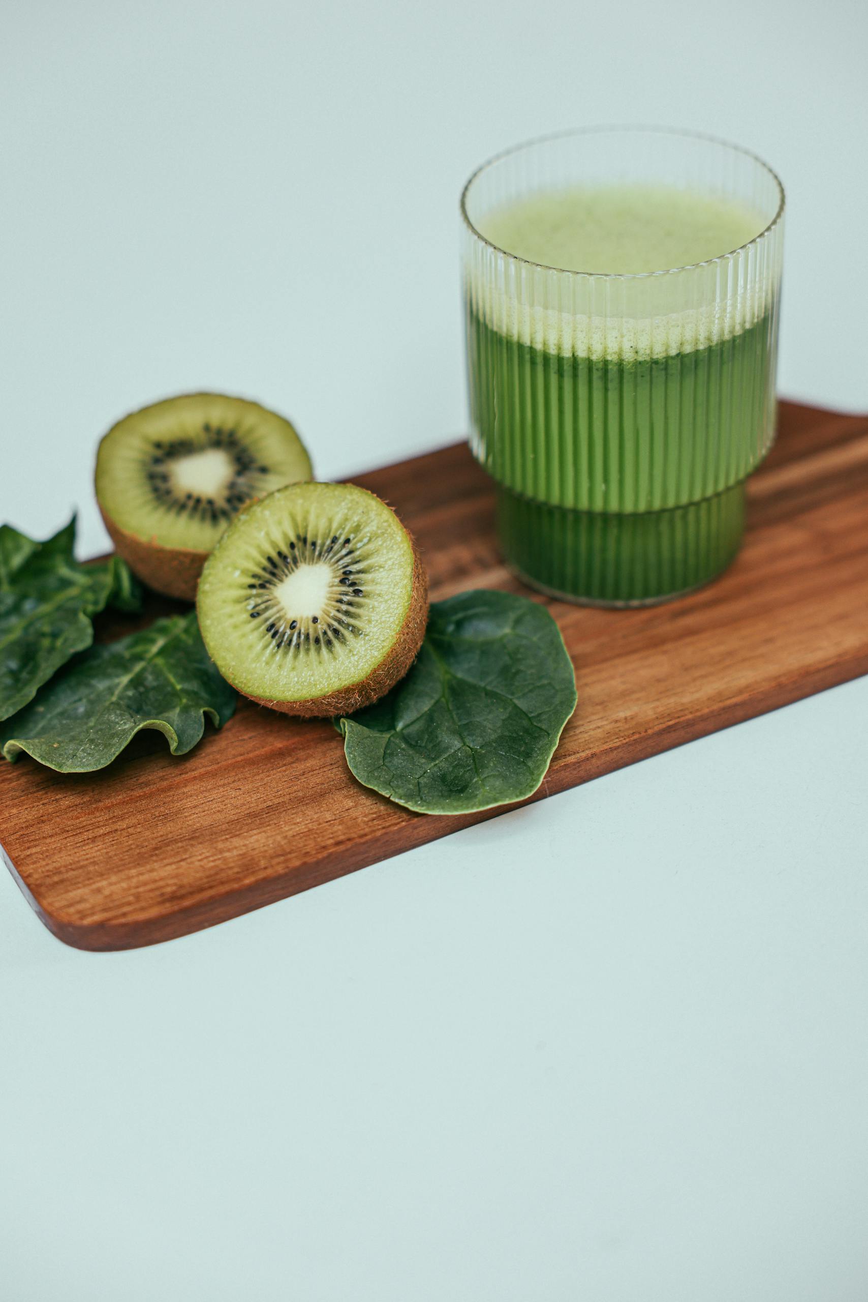 benefits of kiwi: Close-up of a kiwi spinach smoothie in a ribbed glass on a wooden board with sliced kiwi and leaves.