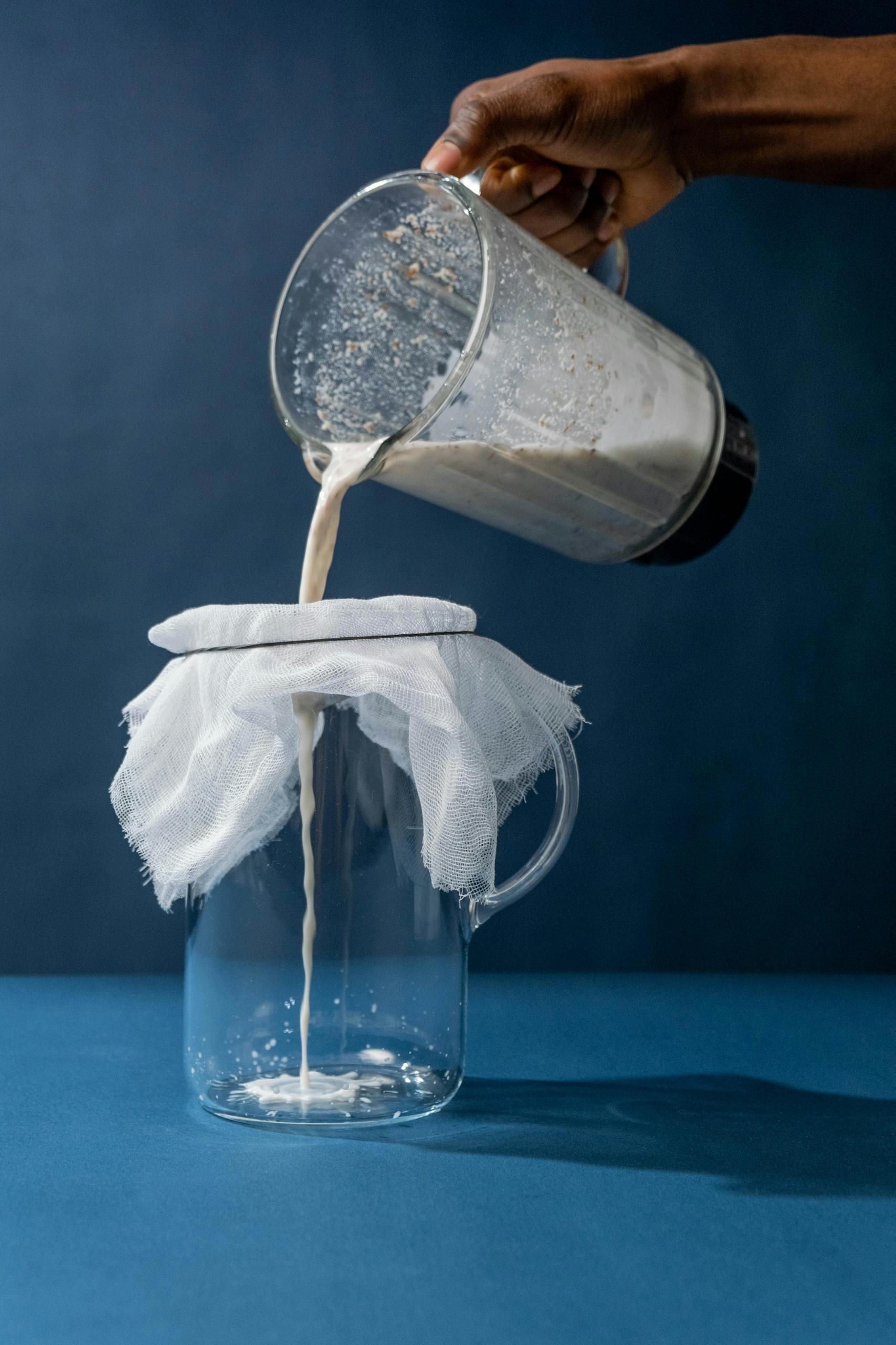 oat milk vs. almond milk vs. soy milk: Hand pours almond milk through cheesecloth into a glass pitcher, showcasing non-dairy preparation.