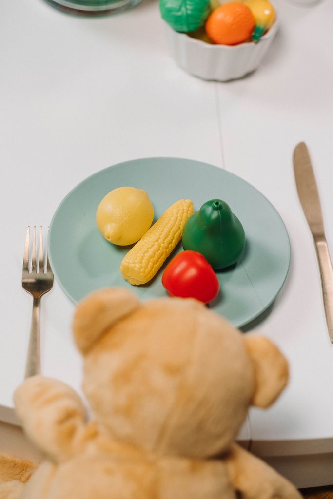 How to Get Your Toddler to Eat Vegetables: Playful scene of a teddy bear with toy vegetables on a dinner plate, viewed from above.