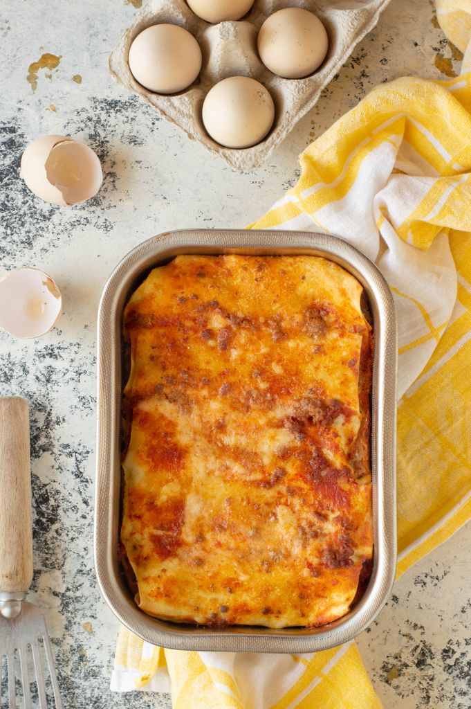 Delicious homemade lasagna in baking dish surrounded by eggs and kitchen utensils.