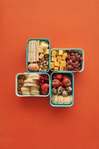 meal prep tips: Top view of four lunch boxes with fruits, cheese, and snacks on orange background.
