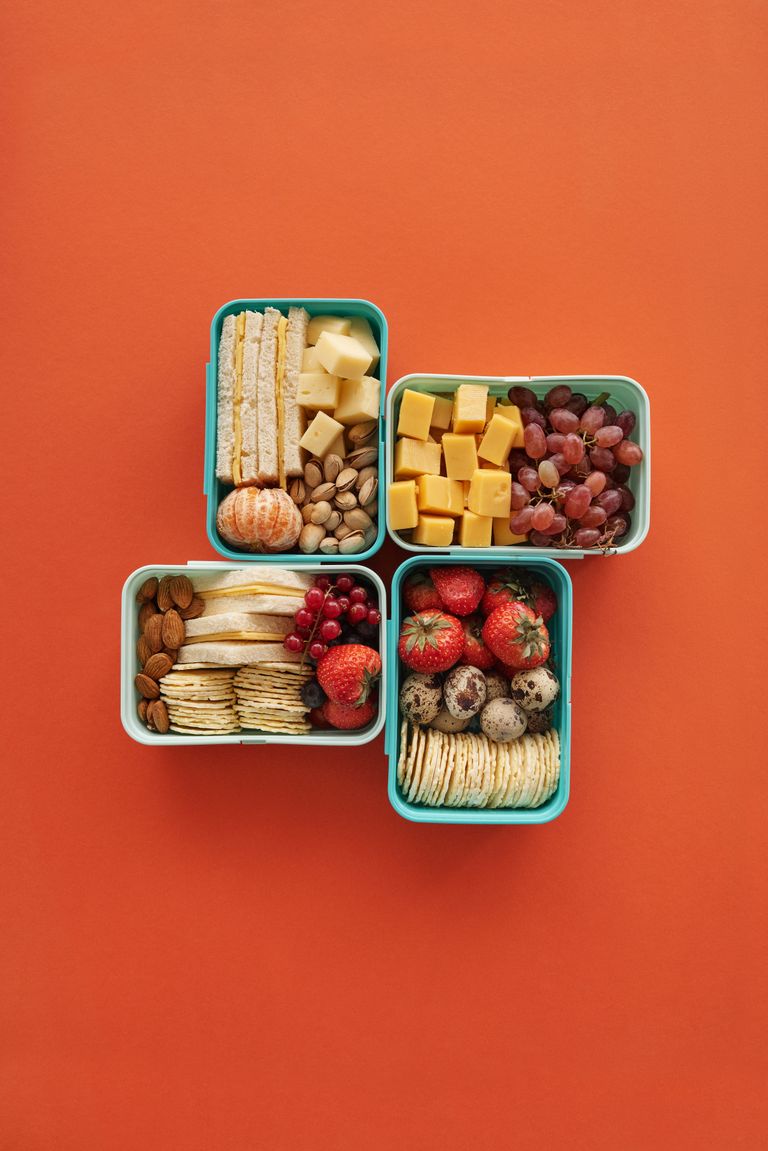 meal prep tips: Top view of four lunch boxes with fruits, cheese, and snacks on orange background.